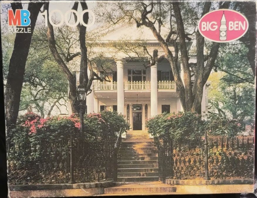 A stately white antebellum-style mansion framed by large trees and a wrought iron fence.