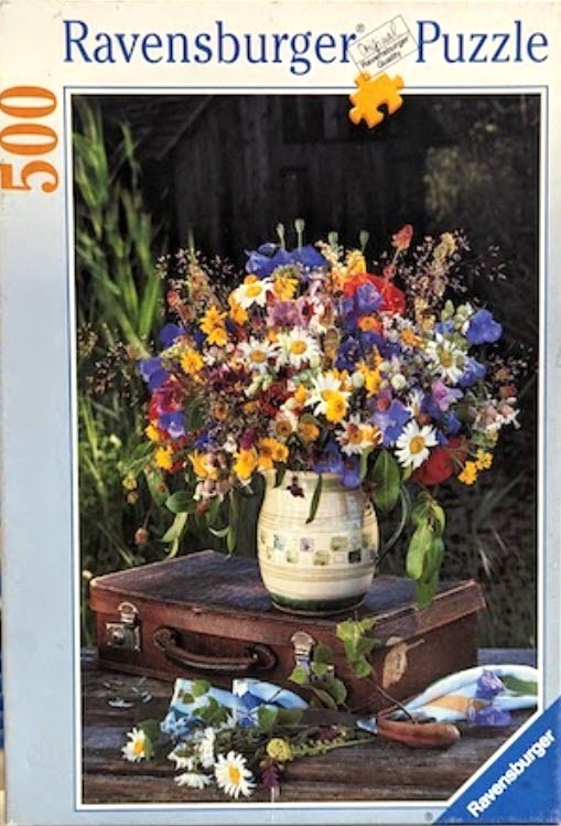 A lush, colorful bouquet of wildflowers in a rustic jug sitting atop an old leather suitcase.