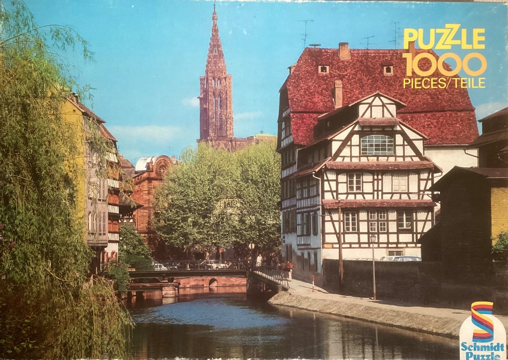 A sunny European canal scene with traditional half-timbered houses and a tall gothic cathedral spire in the background.