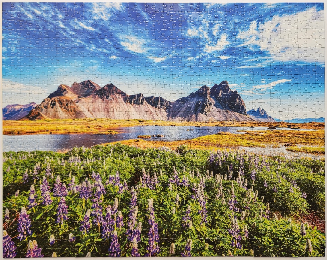 A panoramic landscape featuring purple lupines in the foreground, a calm lake, and sharp, jagged mountains under a blue sky.