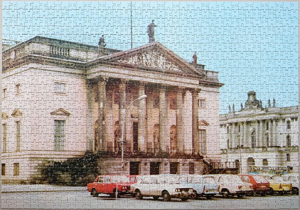 Historic neo-classical stone building with a line of vintage cars parked in front under a clear sky.