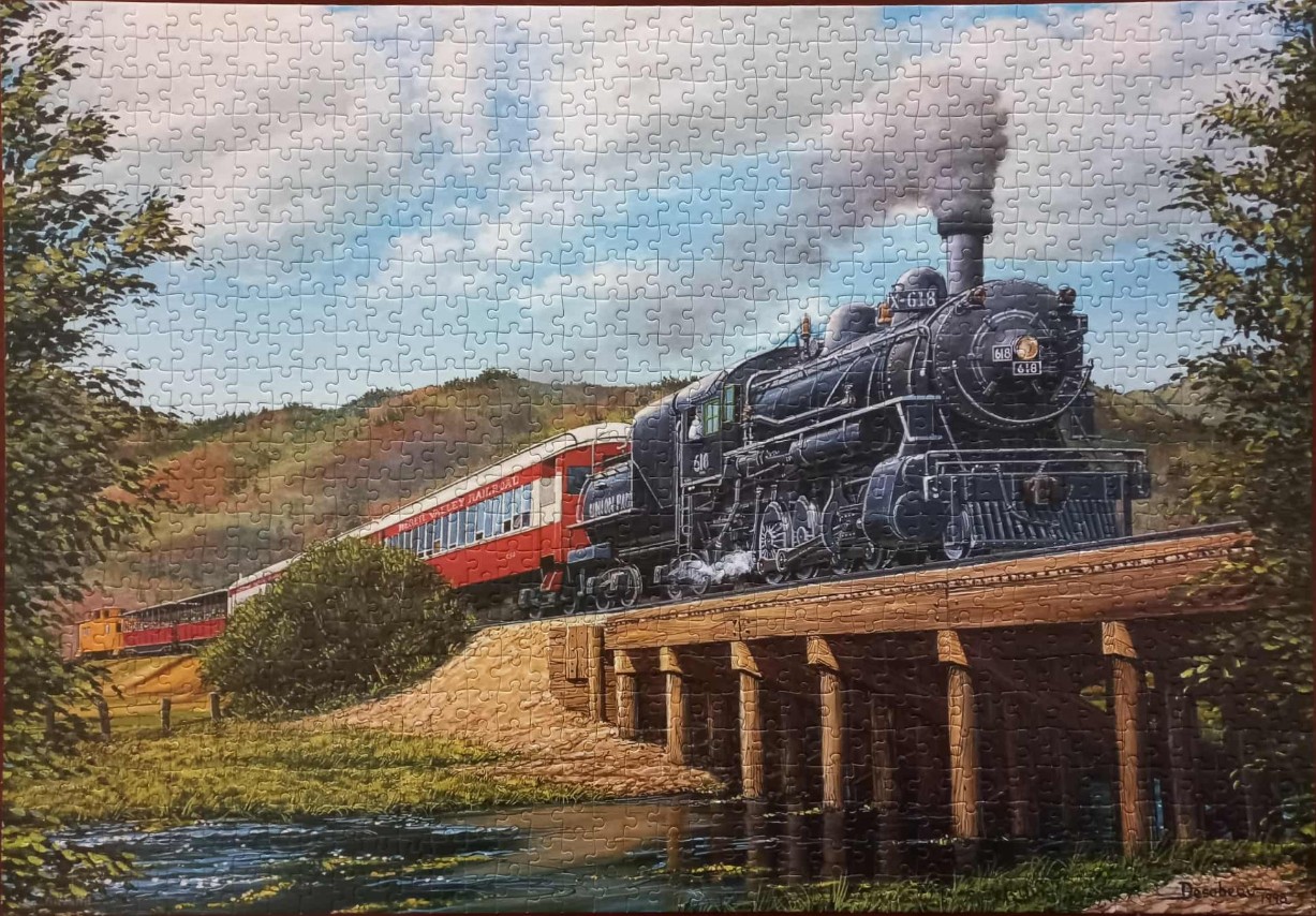A vintage steam locomotive crossing a wooden trestle bridge over a stream in a rural landscape.
