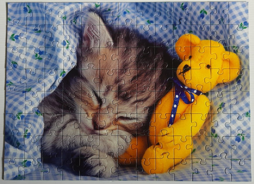 A grey tabby kitten sleeping next to a bright yellow teddy bear on a blue and white gingham blanket.