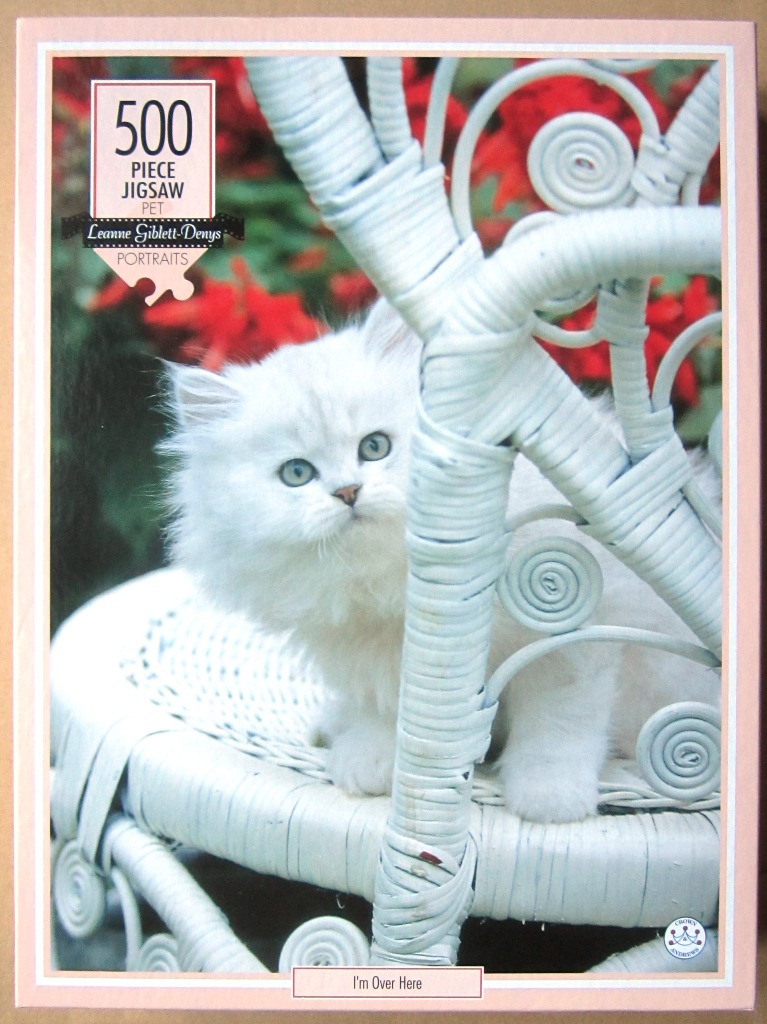 A fluffy white kitten peering out from behind a white wicker chair against a background of red flowers.