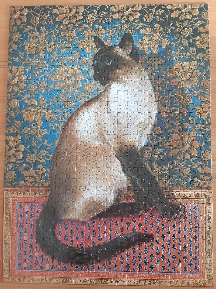 A Siamese cat sits elegantly against a blue floral backdrop with an ornate red and blue patterned rug in the foreground.