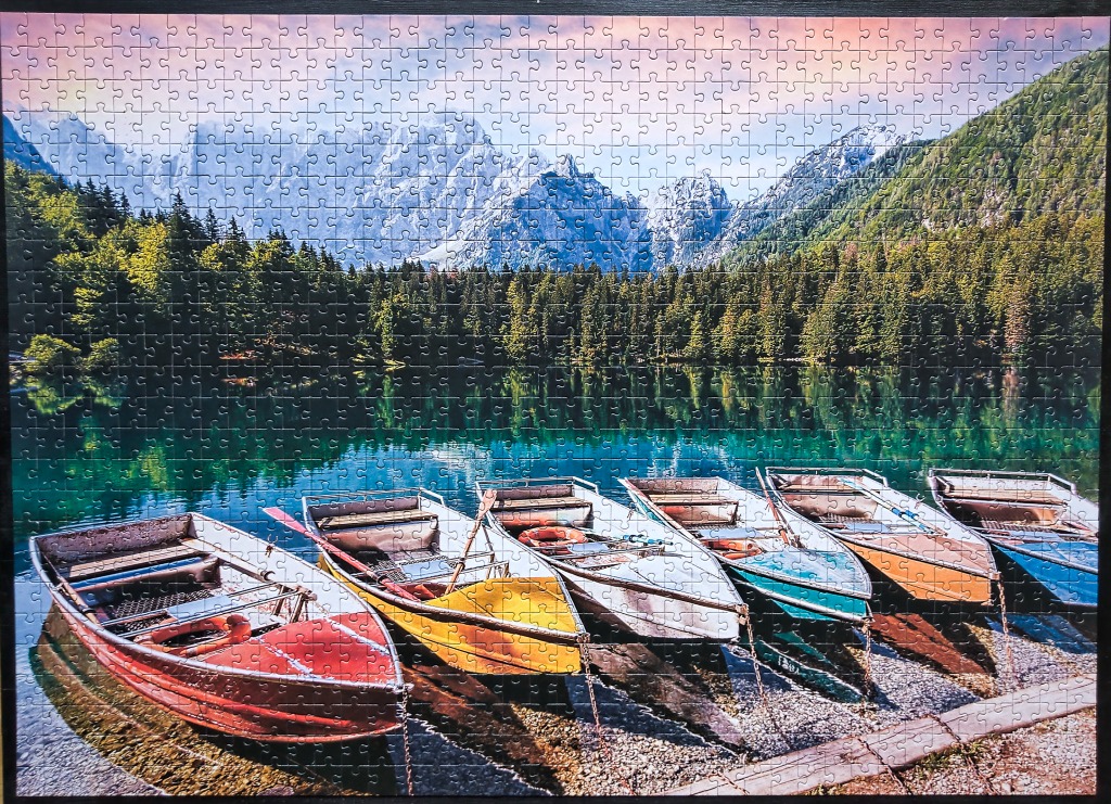 A row of colorful rowboats on the shore of a calm mountain lake with forested slopes and jagged peaks in the background.