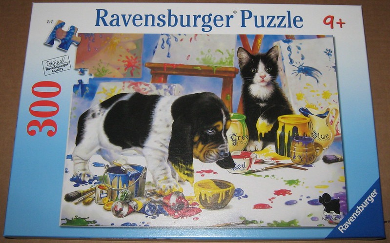 A Basset Hound puppy and a black and white kitten sitting in a messy art studio surrounded by spilled paint and brushes.
