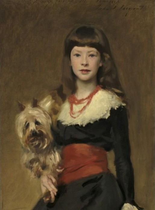 A young girl with long brown hair and bangs holding a small, shaggy terrier dog, wearing a black dress with red accents.