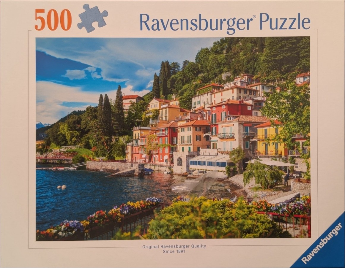 A sunny, colorful Mediterranean village built into a hillside overlooking a blue lake with flowers in the foreground.
