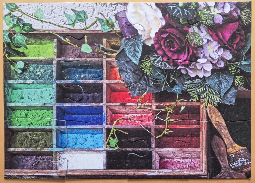 A wooden printer's tray filled with vibrant colorful artist pigments, decorated with flowers and an old paint brush.