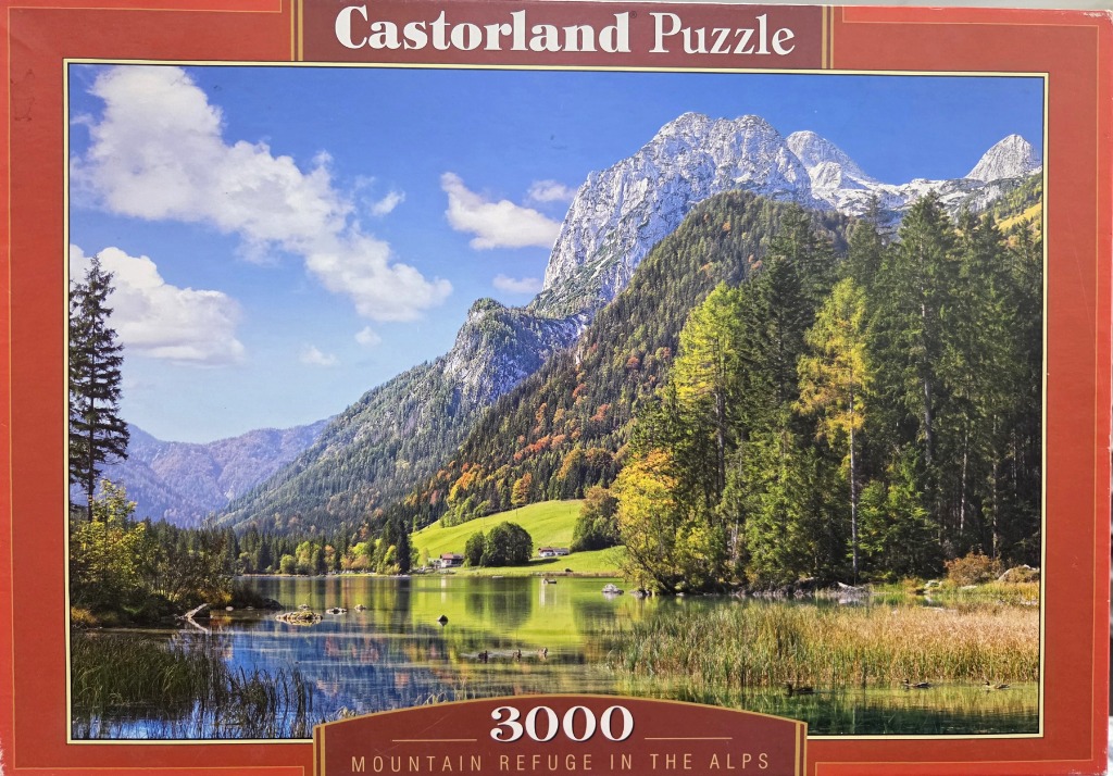 A scenic photograph of a calm lake reflecting rugged, snow-capped Alpine mountains and lush, forested slopes.