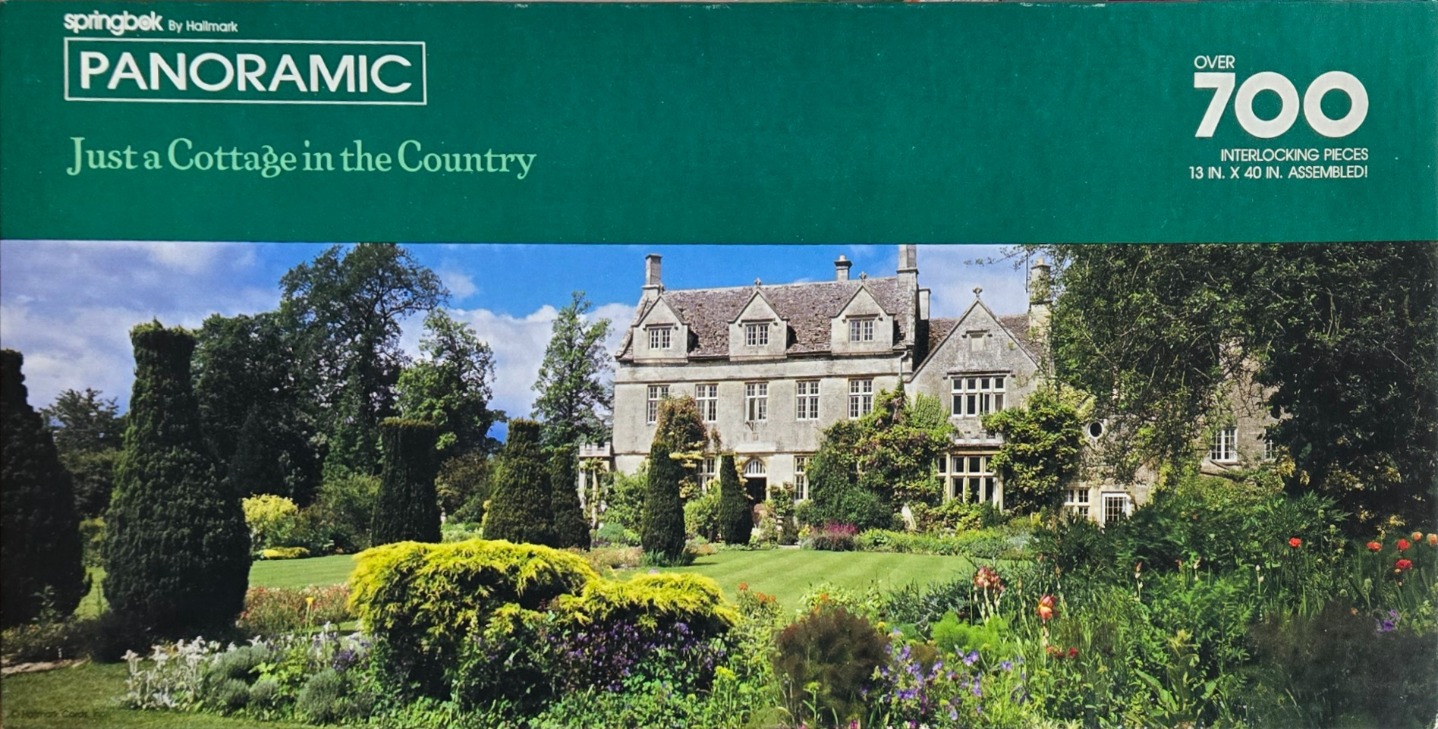 A panoramic photograph of a large, historic stone cottage surrounded by lush, manicured English gardens under a blue sky.