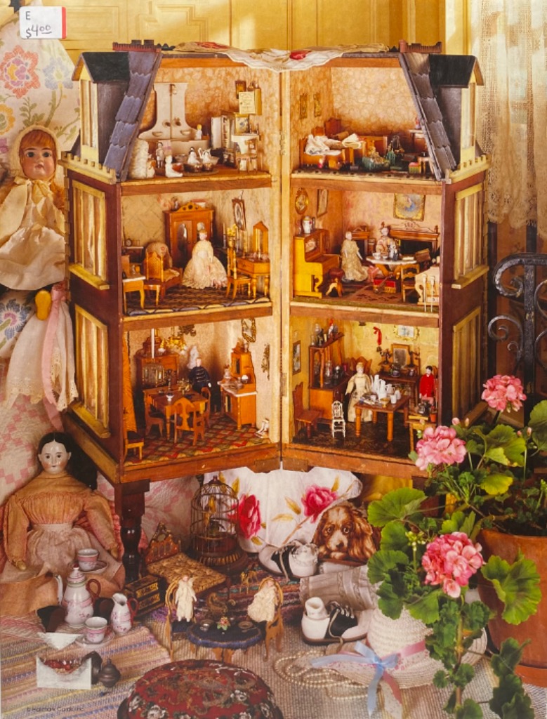 A vintage-style dollhouse open to reveal four furnished rooms, surrounded by dolls, toys, and a potted geranium plant.