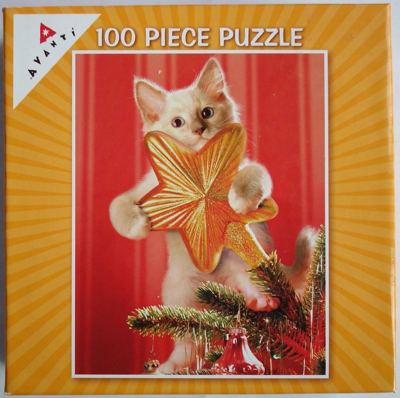 A small white kitten holding a large, shiny golden star ornament on top of a Christmas tree against a red background.