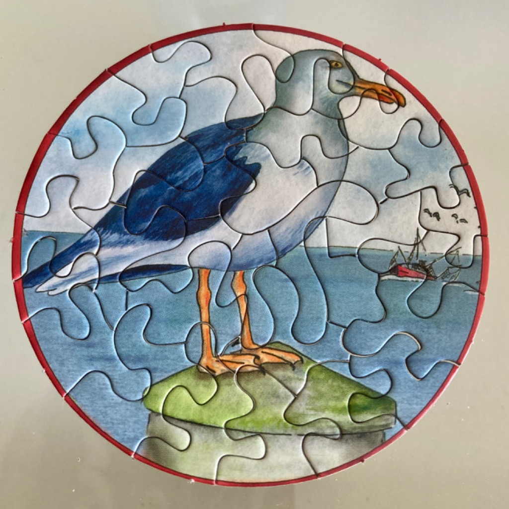 A circular illustration of a seagull standing on a green piling with a small fishing boat in the background.