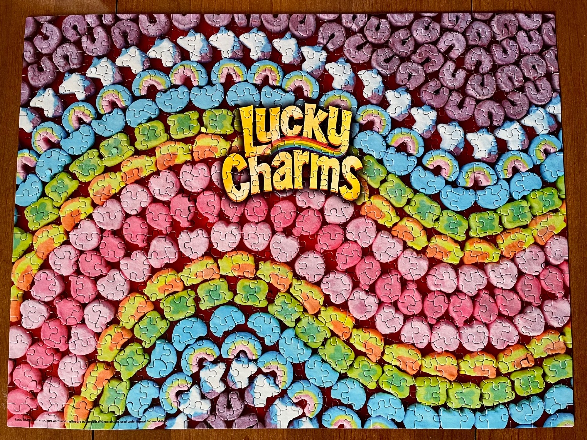 A pattern of colorful cereal marshmallow shapes arranged in concentric rainbow arcs with a brand logo in the center.
