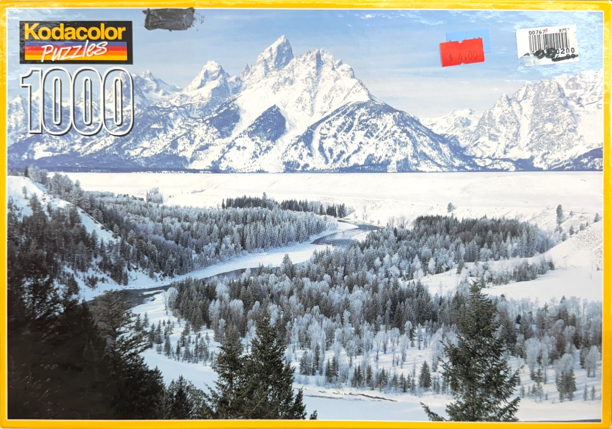 A panoramic winter landscape featuring the jagged, snow-capped Teton mountain range towering over a frozen river valley.