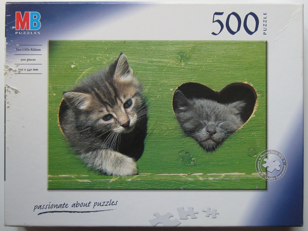 Two kittens peaking through cutouts in a green wooden board, one round and one heart-shaped.