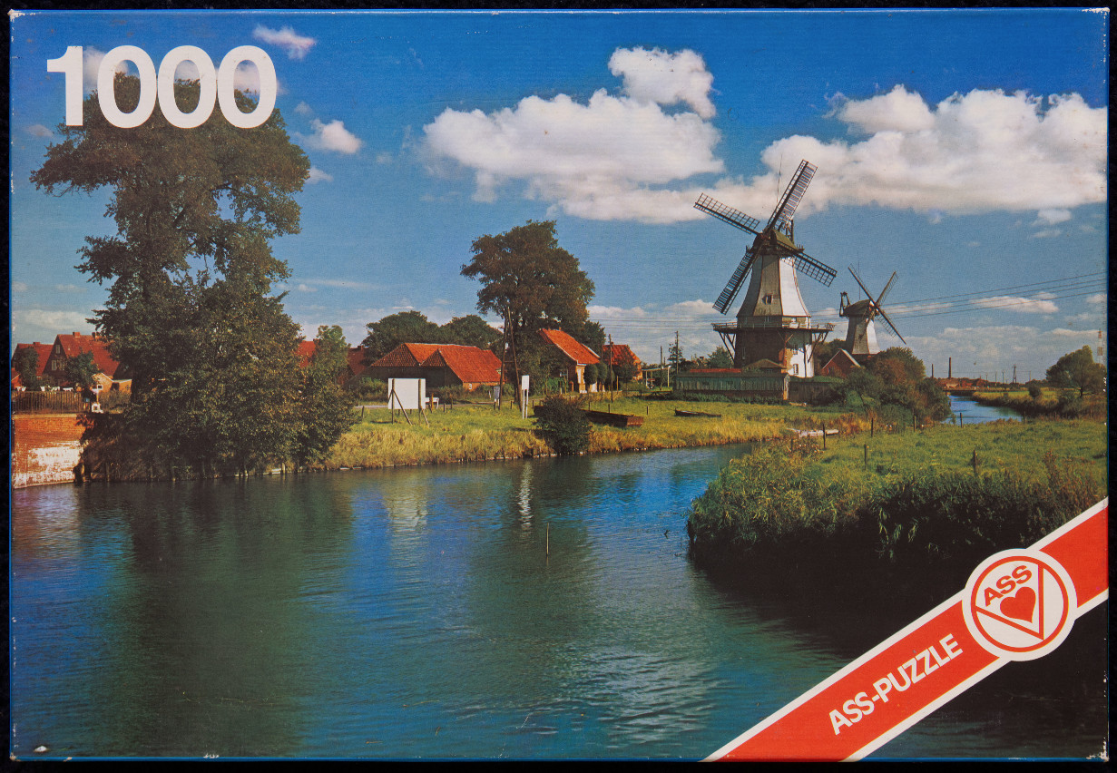 A scenic view of two windmills along a canal in a rural Dutch-style landscape under a blue sky with white clouds.