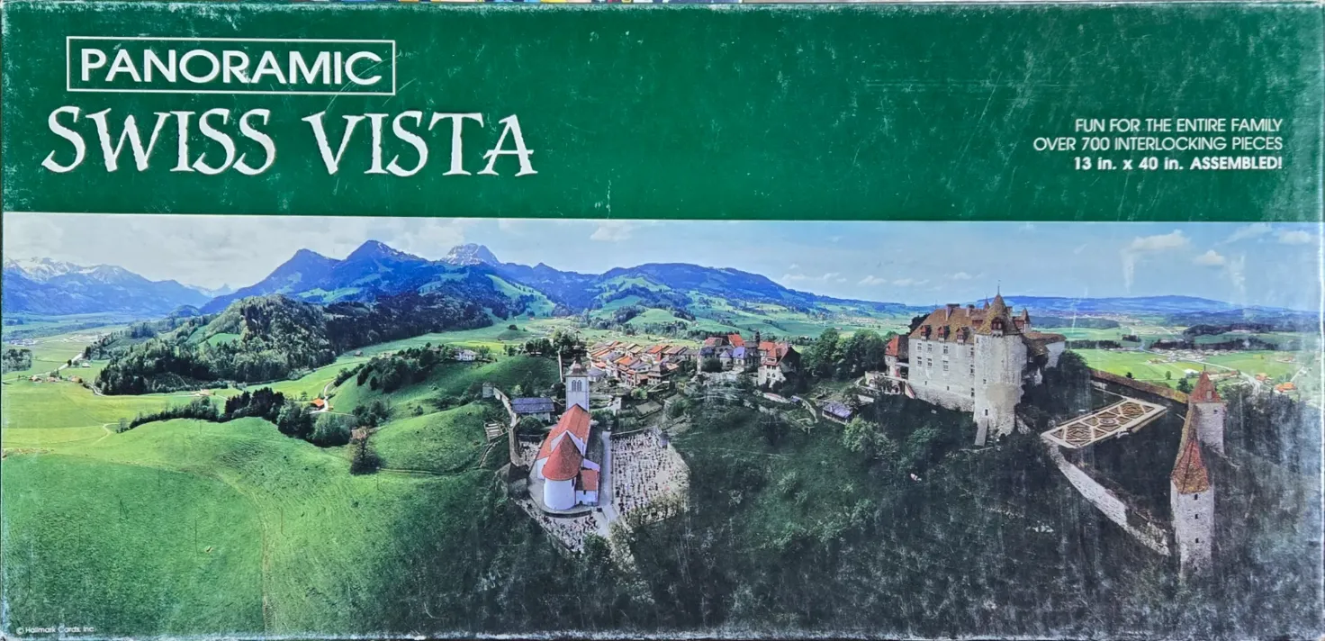 Panoramic view of a medieval Swiss village and castle set against rolling green hills and distant snow-capped mountains.