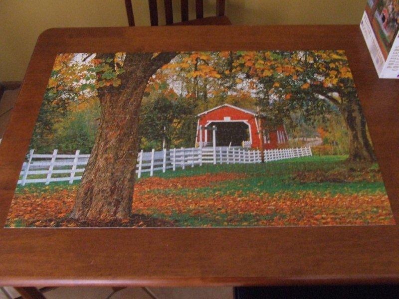 A red covered bridge surrounded by autumn trees and a white fence in a rural landscape.