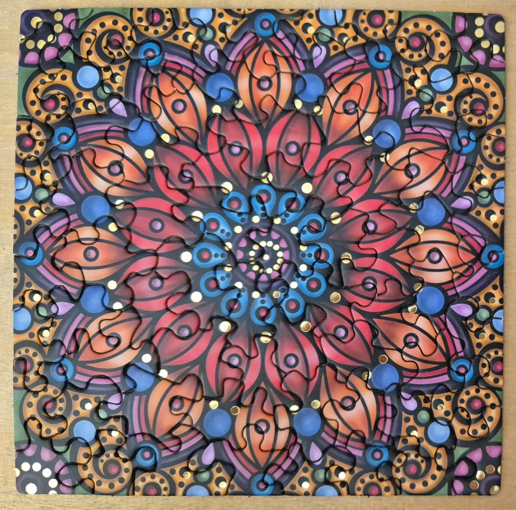 A square, symmetrical mandala-style illustration featuring concentric layers of petals, spirals, and golden dots.