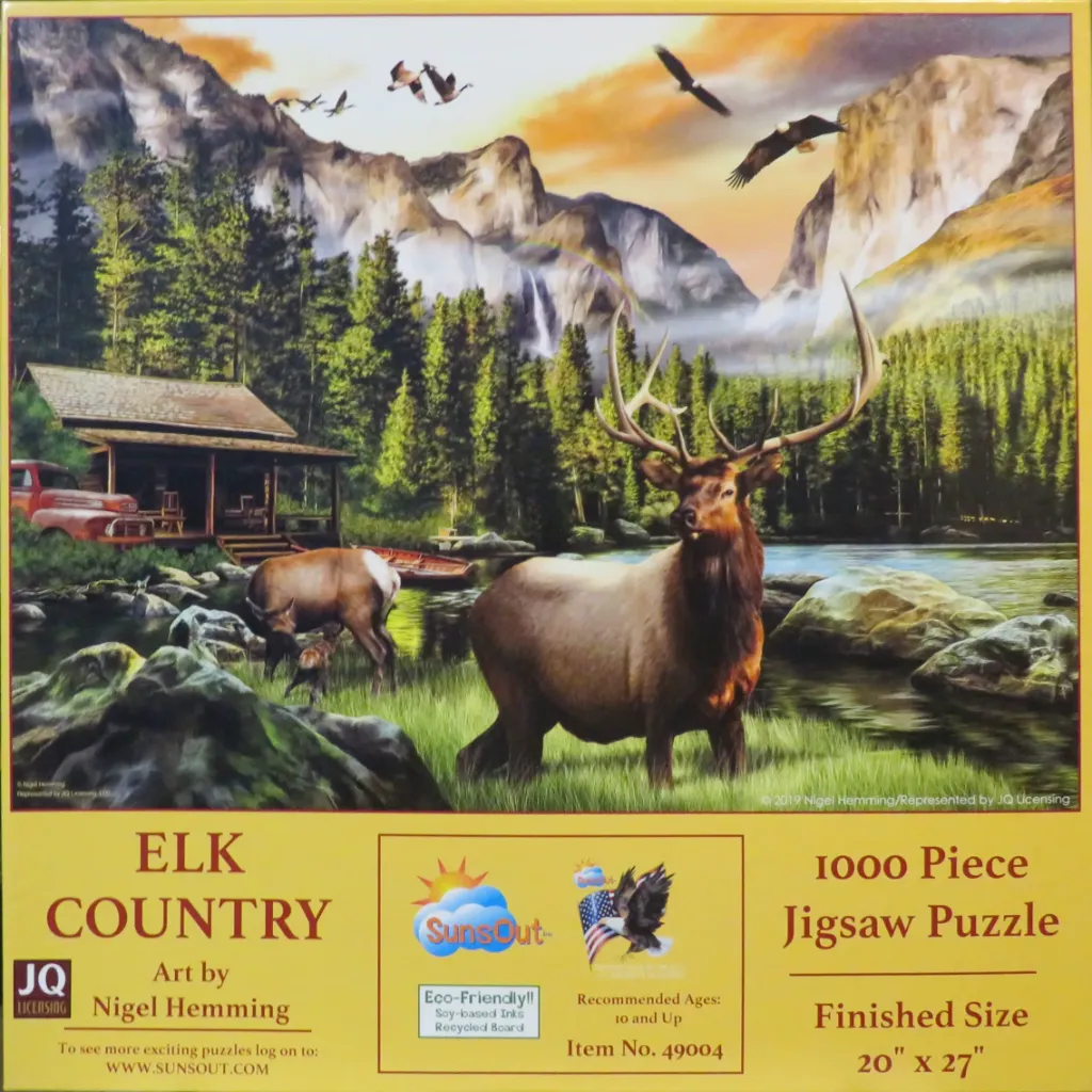 A majestic elk stands in a lush green meadow before a lake and mountain landscape.