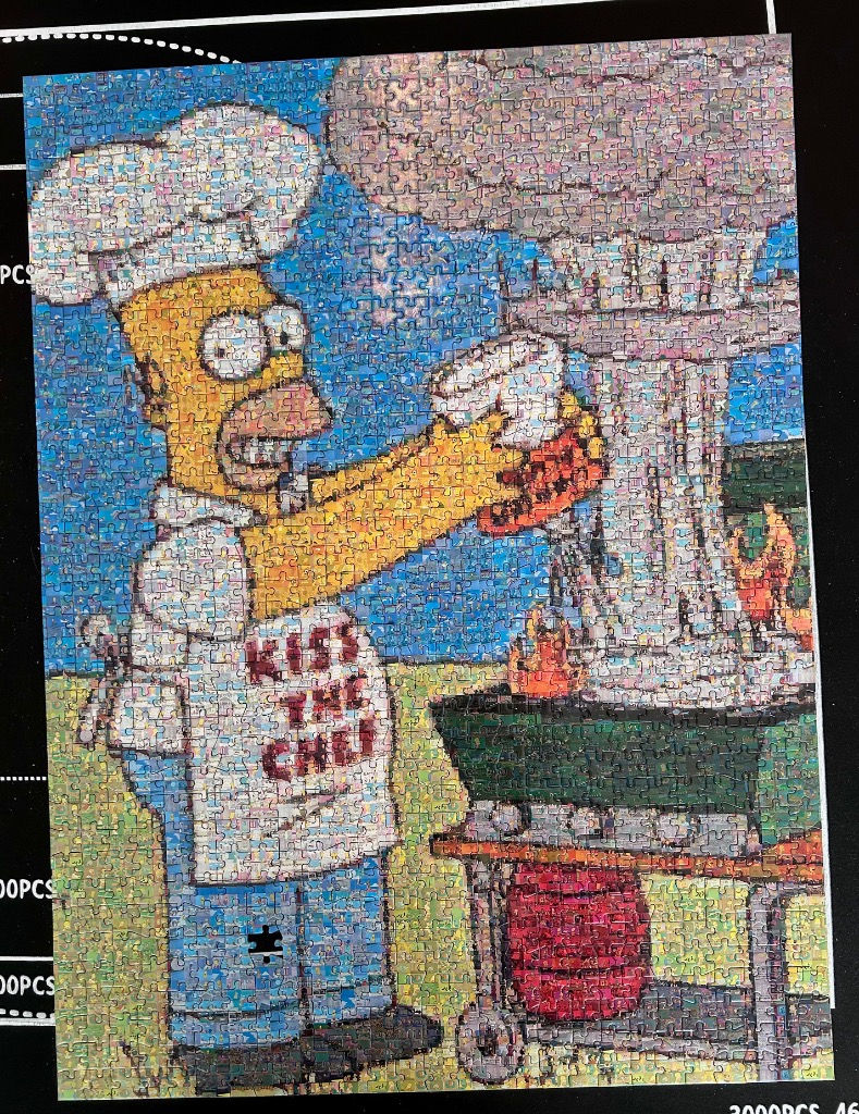 Homer Simpson wearing a chef's hat and a Kiss the Chef apron while grilling outdoors.