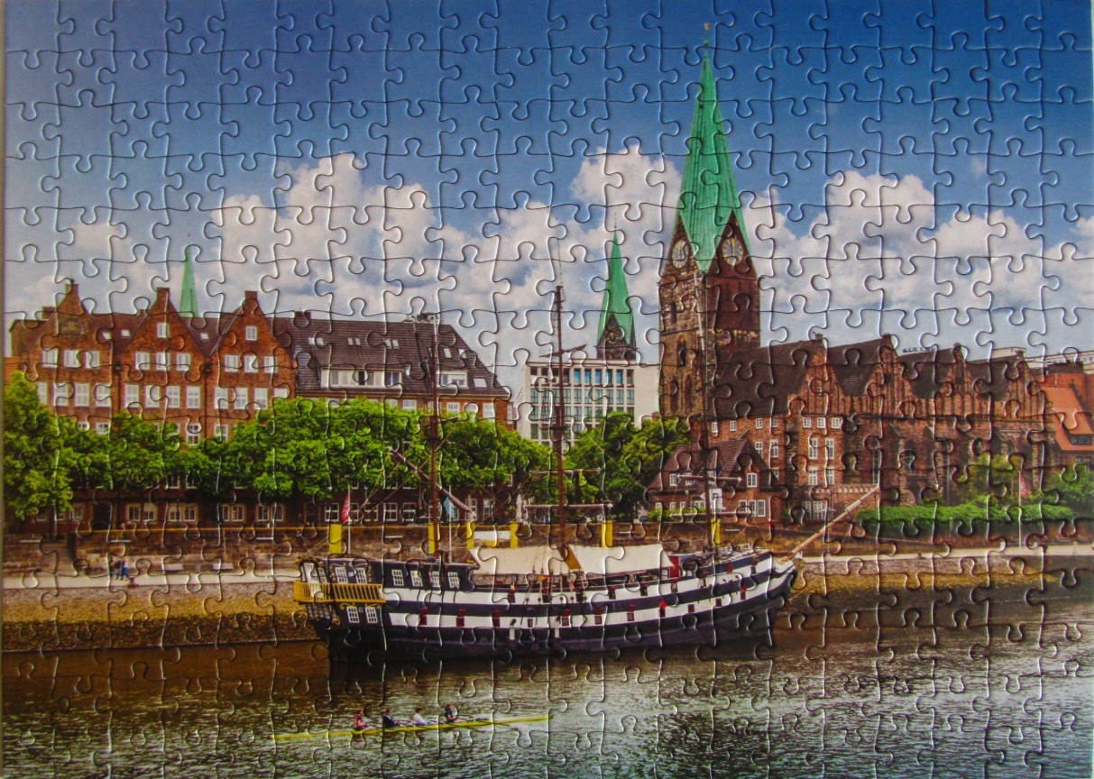 A scenic view of a historical sailing ship docked on a river in front of an old European city with church spires.
