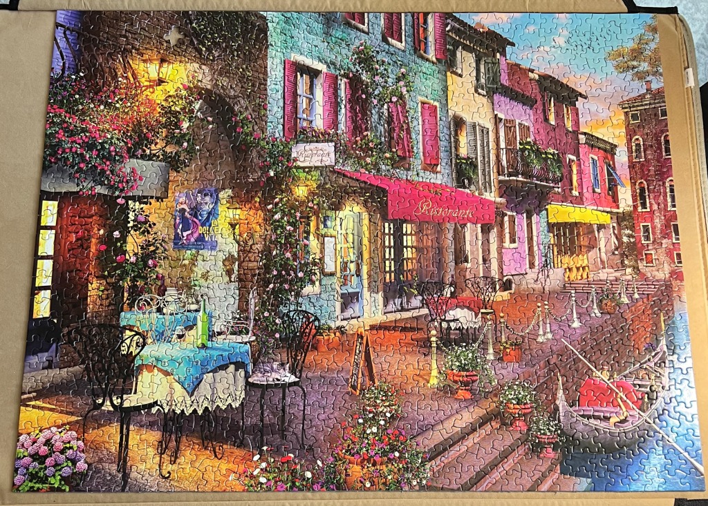 A charming Italian canal-side restaurant scene with colorful buildings, flowers, and a moored gondola.