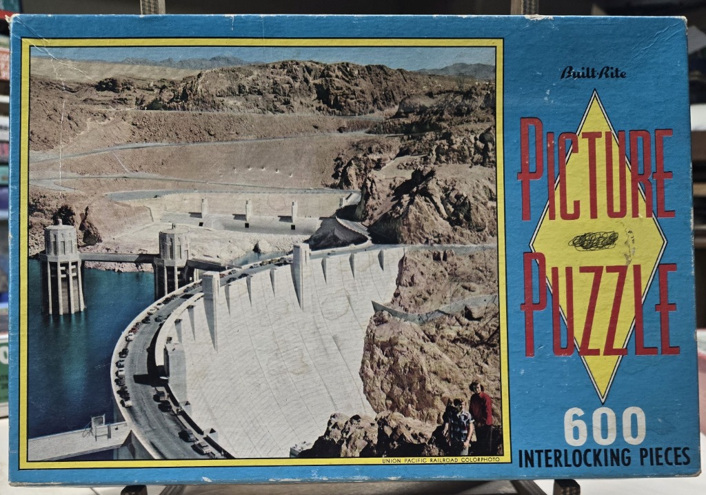 A vintage color photograph of the Hoover Dam with a light blue border and text.
