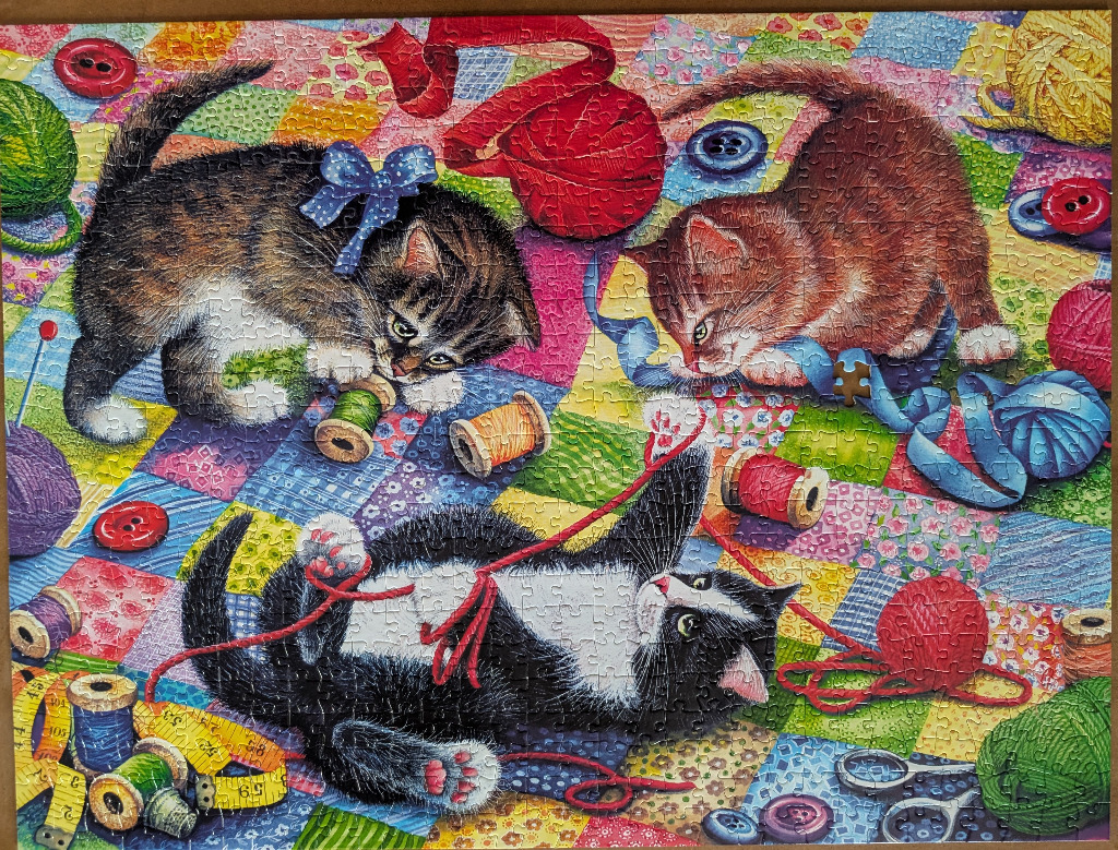 Three playful kittens interacting with yarn, buttons, and sewing supplies on a colorful patchwork quilt background.