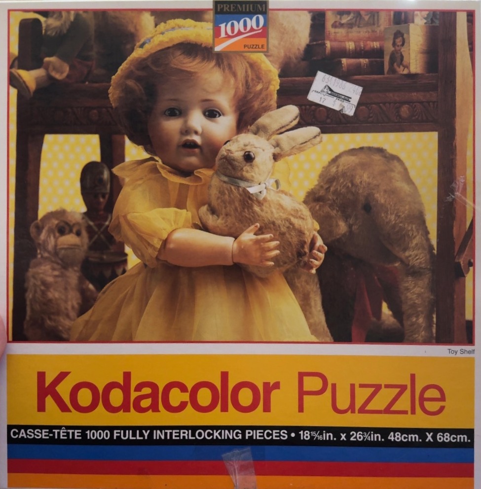 A vintage porcelain doll in a yellow dress holding a plush rabbit, surrounded by other antique toys on a shelf.