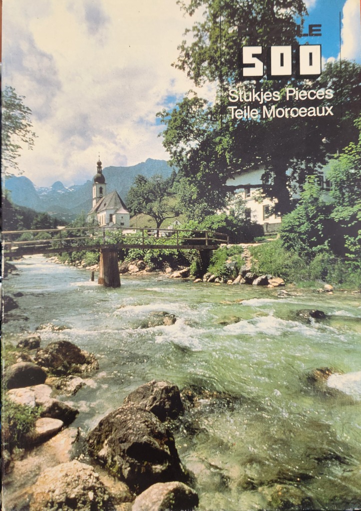 A photograph of a mountain stream flowing toward a small wooden bridge with a traditional alpine church in the background.