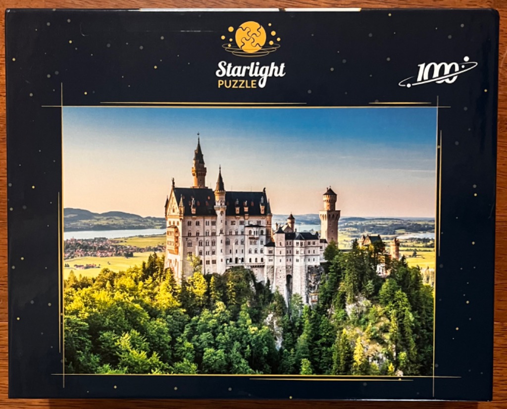 A photograph of Neuschwanstein Castle perched on a forested hilltop under a clear, pale blue sky at sunset.