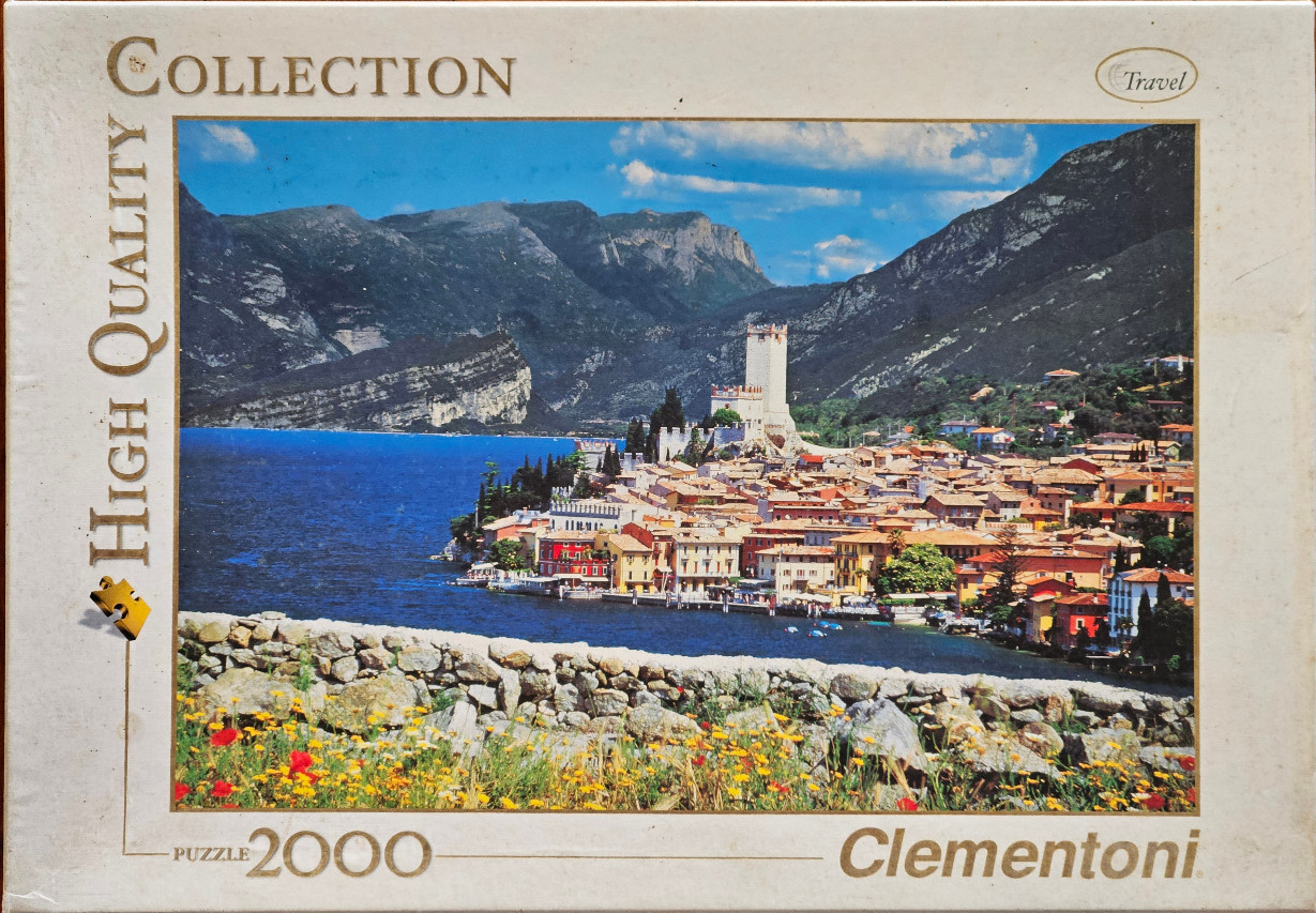 Panoramic photograph of Malcesine on Lake Garda with a medieval castle, mountains, and a stone wall with wildflowers.