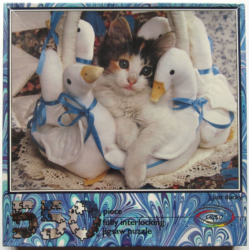 A small calico kitten nestled inside a wicker basket with several plush white duck toys with blue ribbons.