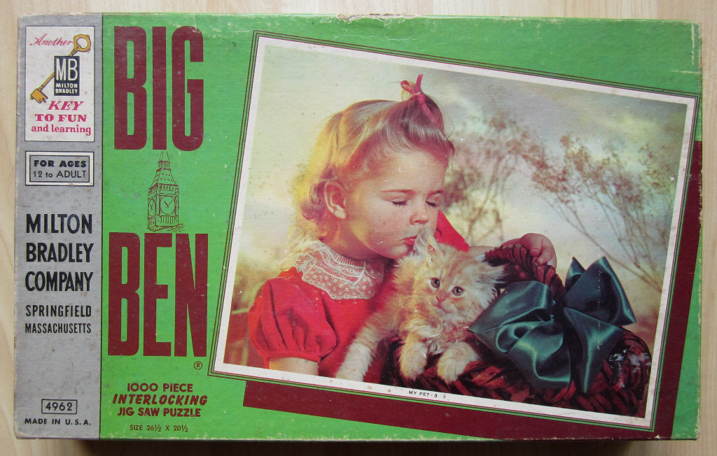 A young girl with blonde hair in a red dress kisses a fluffy ginger kitten sitting in a basket with a large green bow.