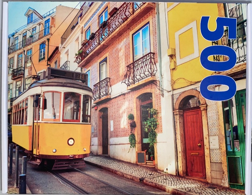 A classic yellow tram travels along a cobblestone street in a historic European city lined with colourful tiled buildings.