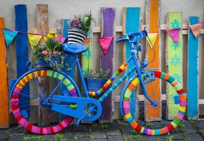A blue bicycle decorated with colorful yarn, parked against a vibrant, multi-colored wooden picket fence.