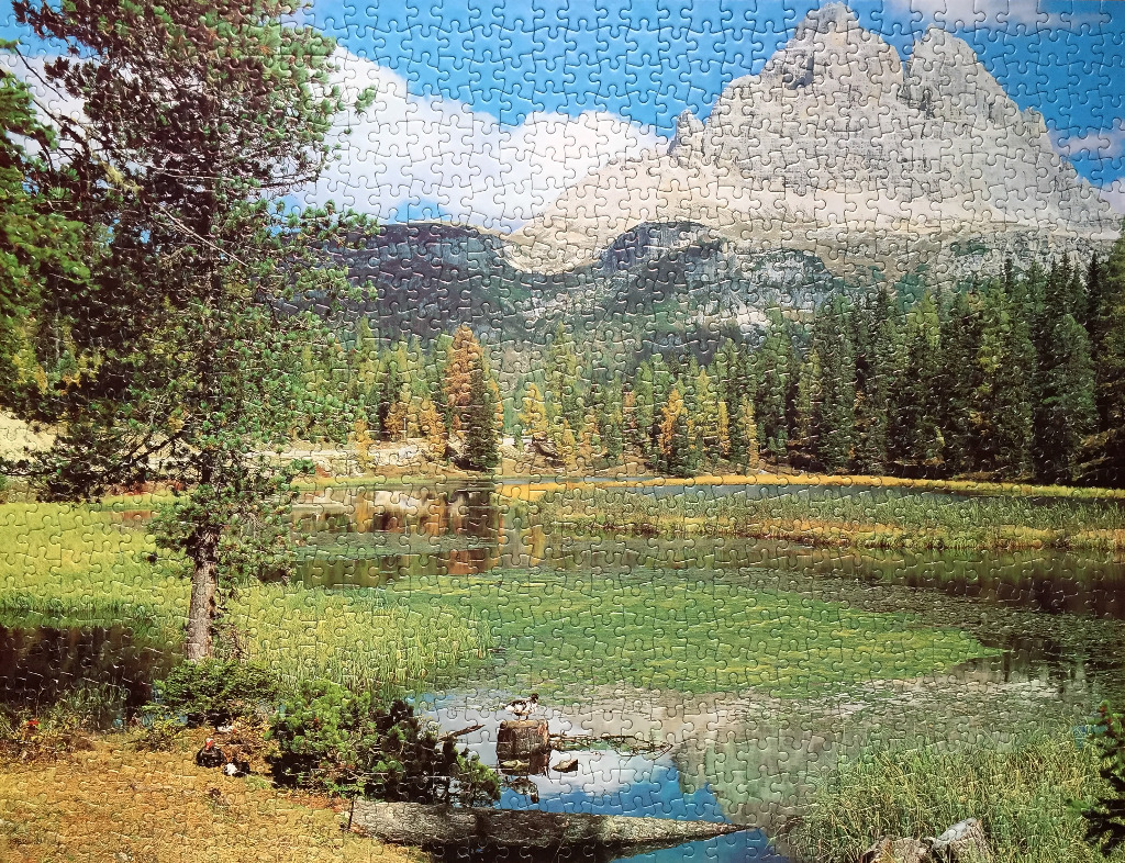 A peaceful alpine scene with a lake, pine trees, and jagged mountain peaks under a blue sky.