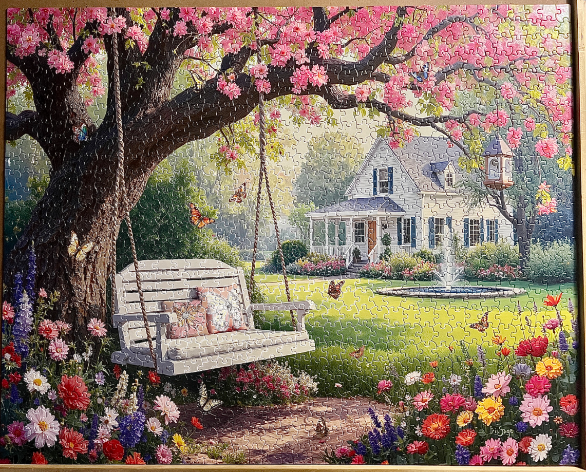 A white garden swing beneath a blooming pink tree in front of a cottage with a fountain.