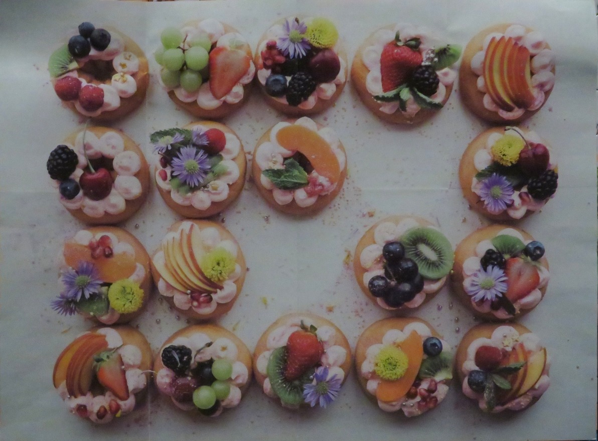A square grid of eighteen decorated donuts topped with colorful fresh fruit, berries, and edible flowers.