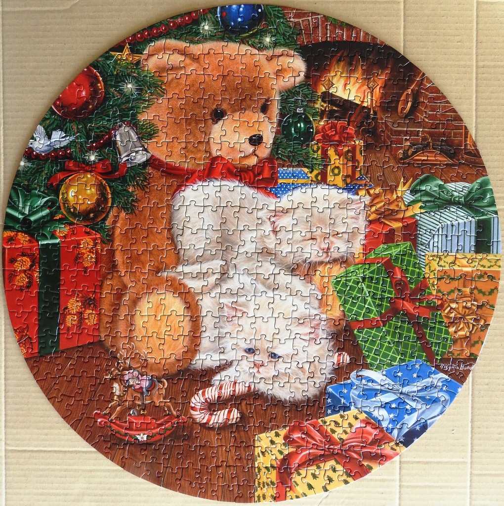 A round illustration featuring a teddy bear and two white kittens sitting by a fireplace with Christmas gifts.