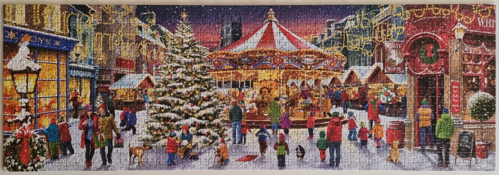 A panoramic Christmas market scene featuring a carousel, a large decorated tree, and people shopping in a snowy town square.