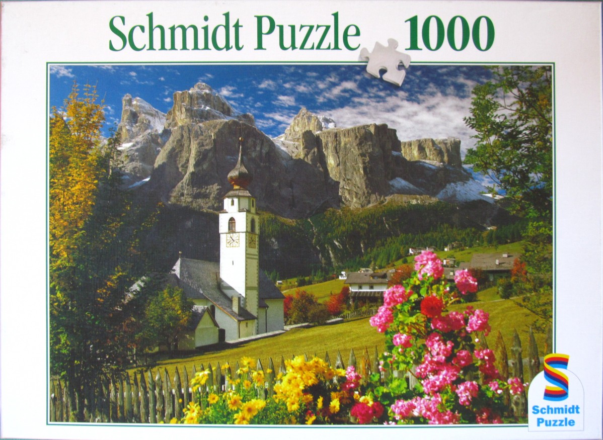 A scenic alpine view featuring a church with a tall steeple in front of jagged mountains and a field of flowers.