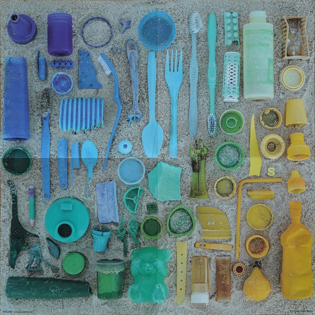 An overhead collection of discarded plastic items, sorted by color, lying on sandy ground.