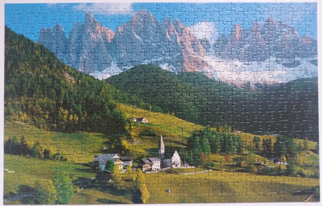 A small village with a church nestled in a green valley before the jagged, snow-dusted peaks of the Dolomites.