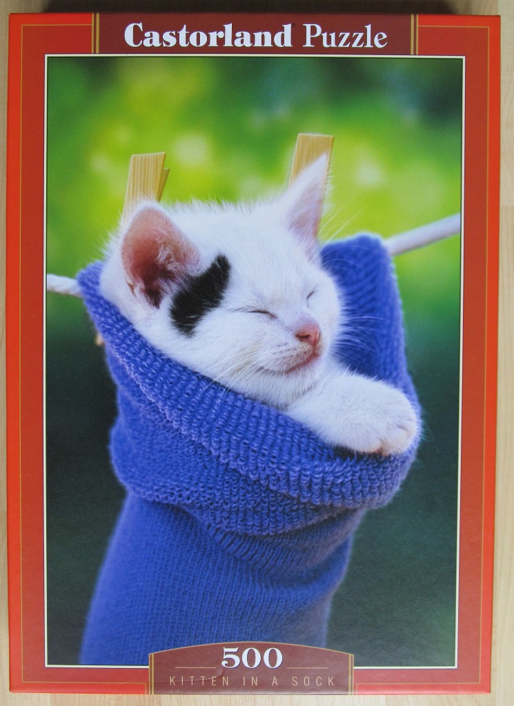 A white kitten with a black ear patch sleeping inside a bright blue knit sock hanging from a clothesline.