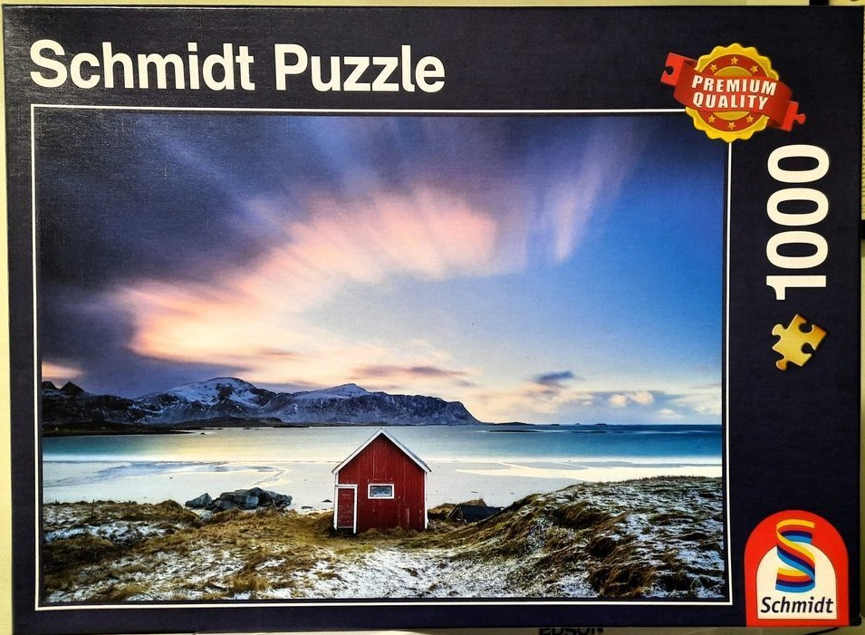 A red wooden cabin on a snowy beach against a sunset sky and mountains in Norway.